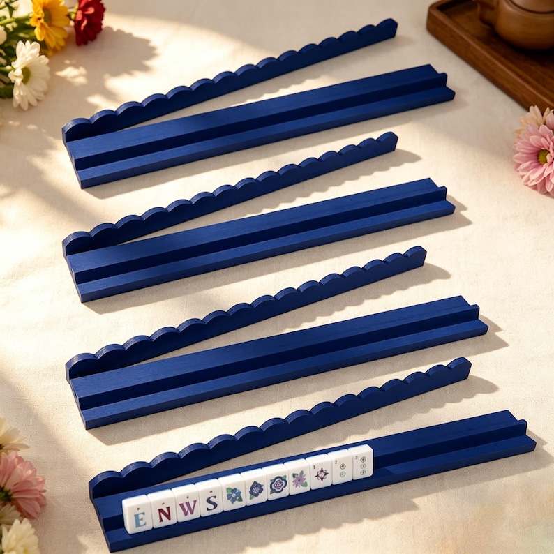 May include: A set of four long, dark blue wooden racks with scalloped top edges and two parallel horizontal grooves for holding game tiles. One rack contains a row of white rectangular tiles featuring letters and floral patterns.