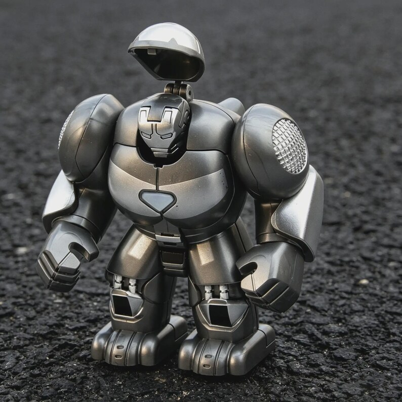 以下が含まれることがあります： 開閉可能なヘルメットが付いた、シルバーとグレーのロボットのおもちゃ。丸みを帯びた肩、胸部プレート、関節式の腕と脚を備えた詳細なデザインです。暗く、質感のある表面に立っています。