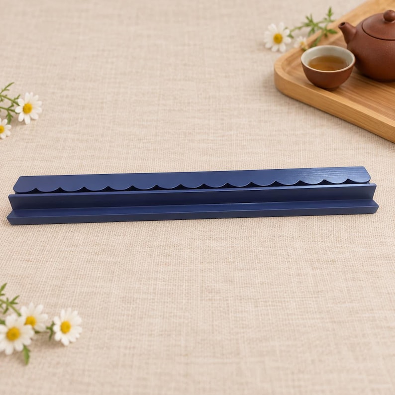 May include: A long, narrow, dark blue wooden display shelf featuring a decorative scalloped top edge. The shelf has a flat base and a recessed groove designed for holding items upright, showcasing a smooth, matte finish across its entire length.