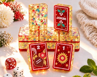May include: A set of rectangular game tiles with gold glitter and red resin, featuring printed designs including a Joker card, a number 2 with floral motifs, and a number 1 with a sunburst pattern, accompanied by two red and white dice.