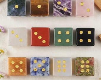 May include: A collection of twelve square dice crafted from various polished stones, including moss agate, tiger's eye, amethyst, rose quartz, and others. Each die features gold-colored circular pips indicating values from one to six.