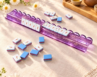 May include: A purple translucent scalloped Mahjong tile rack holding a row of white tiles with cocktail illustrations, accompanied by several loose white tiles with cocktail graphics and solid blue tiles scattered on a surface.