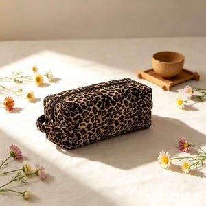 May include: A rectangular quilted pouch featuring a black and tan leopard print pattern. The pouch has a black zipper closure along the top and a small fabric handle on the side. The exterior fabric has a stitched diamond pattern throughout.