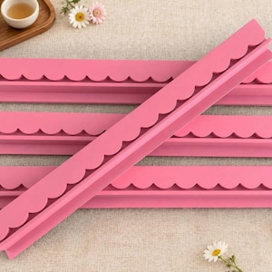 May include: Four long, pink, L-shaped shelf strips featuring a decorative scalloped edge along the top. The strips are arranged with three lying horizontally and one placed diagonally across them, showcasing the uniform matte finish.