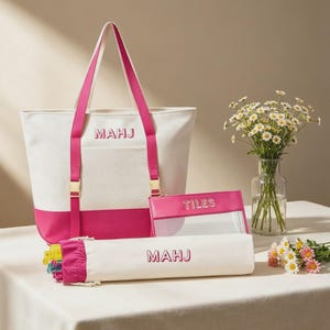 Könnte beinhalten: Eine weiße und pinke Tragetasche aus Segeltuch mit dem Text MAHJ, ein passendes zylindrisches Etui mit dem Text MAHJ und eine rechteckige, transparente Tasche mit pinkem Rand und dem Text TILES. Das Set hat goldfarbene Beschläge und pinke Riemen.