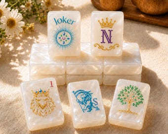May include: A set of rectangular, pearlescent white mahjong tiles arranged in a stack. The top tiles feature a blue Joker design and a gold crown with the letter N. The bottom row displays a gold lion head, a blue lion, and a green tree.