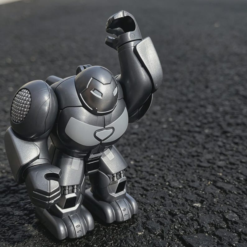 以下が含まれることがあります： 腕を上げたグレーとシルバーのロボットのおもちゃ。 ロボットは丸みを帯びた体、目のスリットのある詳細な頭、関節のある手足を持っています。 おもちゃはプラスチック製で、暗い表面に立っています。