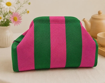 May include: A structured pouch featuring a vibrant knit pattern of alternating vertical stripes in bright magenta and forest green. The bag has a curved top frame closure and a soft, textured fabric construction with a clean, rectangular base.