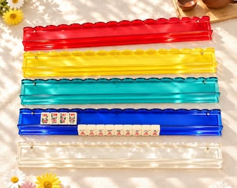 May include: Five long, rectangular acrylic mahjong tile racks in red, yellow, teal, blue, and clear. The blue rack holds twelve white mahjong tiles with red and black Chinese characters and floral designs. Each rack features a scalloped top edge.