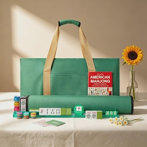Könnte beinhalten: Ein grünes Mahjong-Set bestehend aus einer großen rechteckigen Tragetasche mit beigen Griffen, einer passenden grünen Mattenhülle, einem roten Lehrbuch mit dem Titel American Mahjong, grünen Ablageleisten, bunten runden Chips und Mahjong-Steinen.