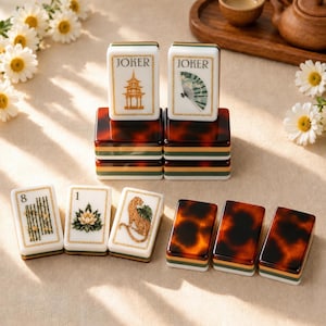 Puede incluir: Un juego de fichas de Mahjong rectangulares con caras blancas e ilustraciones en tono dorado de una pagoda, un abanico, bambú, un loto y un leopardo. Las fichas tienen dorsos con patrón de carey y bases superpuestas en verde, blanco y oro.