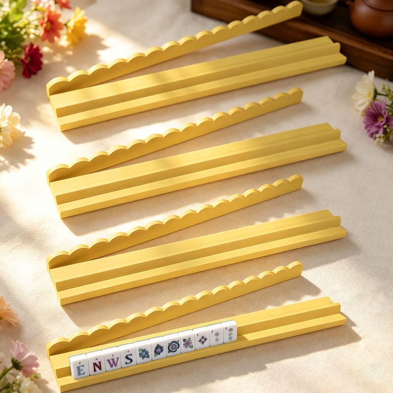 May include: Four yellow wooden tile racks with scalloped top edges and tiered grooves. The bottom rack holds a row of white game tiles featuring letters and floral patterns, including the letters E, N, W, and S printed in black on the tiles.