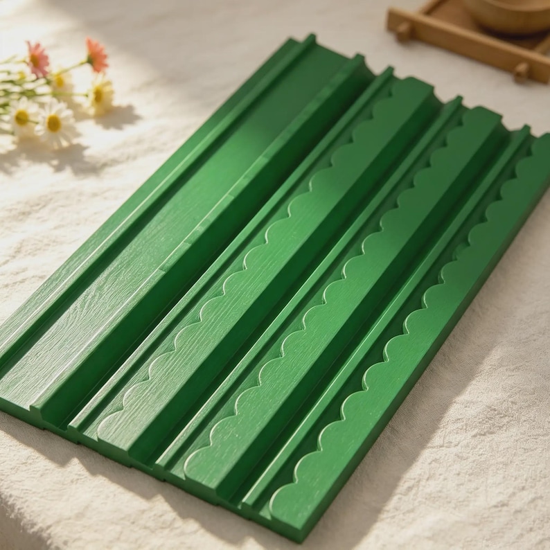 May include: A rectangular green wooden display board featuring three parallel horizontal grooves. The board has a distinct scalloped edge design along the raised sections, providing a textured surface for organizing or showcasing small items.