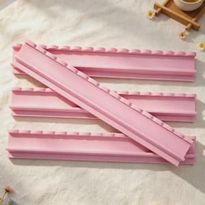 May include: Three light pink silicone molds with a long, rectangular shape and a scalloped top edge. Each mold features a central channel and a flat base, designed for creating long, decorative strips or borders with a consistent pattern.