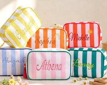 May include: A collection of rectangular zippered pouches featuring vertical stripes in yellow, orange, red, light blue, pink, and teal. Each pouch is embroidered with a name: Charlotte, Sophia, Michelle, Abigail, Athena, and Luxury.