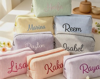 May include: A collection of rectangular zippered pouches featuring vertical pinstripes in various pastel colors. Each pouch is embroidered with a unique name in cursive script, including Haifa, Marina, Reem, Payton, Isabel, Lisa, Kaka, and Raya.