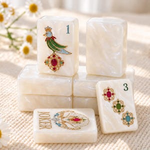 May include: A collection of white pearlescent mahjong tiles. One tile features a bird with a red, green, and blue design and the number 1. Another tile displays three oval gems in red, green, and blue with the number 3. A Joker tile shows a shell design.