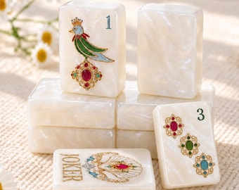 May include: A set of white pearlescent mahjong tiles featuring intricate designs. One tile displays a bird with a crown and a red gemstone, another shows the number 3 with three ornate gemstone motifs, and one is labeled JOKER with a shell design.