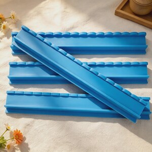 May include: A set of four bright blue silicone molds, each featuring a long, straight channel with a scalloped top edge. These rectangular molds are designed with a flat base and raised sides, presenting a uniform, smooth texture throughout.