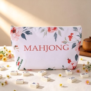 Può includere: Una pochette bianca con cerniera caratterizzata da un motivo floreale con fiori rosa e rossi e foglie verdi. La parola MAHJONG è stampata in grandi lettere maiuscole rosse centrate. La pochette è circondata da tessere del mahjong bianche.