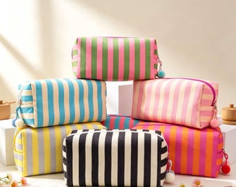 May include: A collection of rectangular zippered pouches featuring vertical stripes in various color combinations including pink and green, blue and cream, pink and cream, yellow and gray, black and white, and orange and pink, each with a small pom-pom.