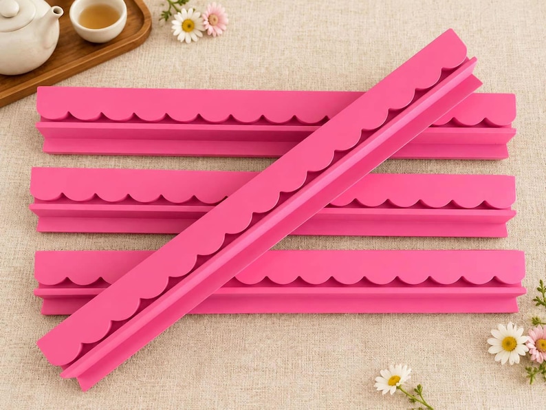 May include: Four bright pink silicone molds, each featuring a long, straight edge with a repeating scalloped pattern along one side. The molds are rectangular in shape and are arranged in a stacked formation with one piece placed diagonally across the others.