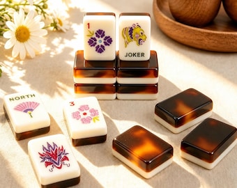 May include: A set of rectangular mahjong tiles featuring a white face with colorful illustrations and text including 'NORTH', 'JOKER', and numbers, paired with a dark brown tortoiseshell-patterned back. The tiles are arranged in stacks and rows.