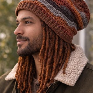 Könnte beinhalten: Ein mehrfarbiger, gestreifter Häkelbeanie in Braun-, Orange- und Grautönen. Der Hut wird von einer Person mit langen, rotbraunen Dreadlocks und einer grünen Jacke mit cremefarbenem Shearling-Kragen getragen.