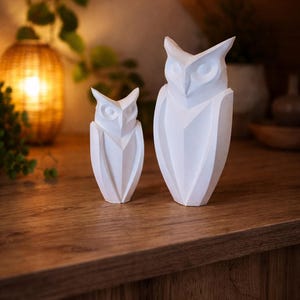 Könnte beinhalten: Zwei weiße Eulenfiguren in verschiedenen Größen, platziert auf einer Holzoberfläche. Die Eulen haben ein geometrisches Design mit spitzen Ohren und einem dreieckigen Schnabel. Der Hintergrund umfasst warmes Licht und Grün.