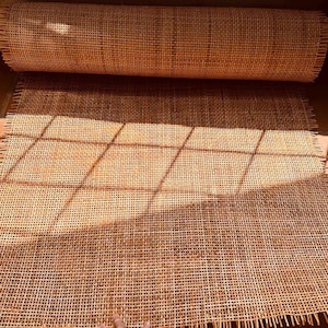 May include: A roll of light brown woven rattan cane webbing featuring a consistent square grid pattern. The material is flexible and displays a natural tan color with visible fibers throughout the mesh, showing frayed edges along the sides.
