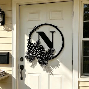 Puede incluir: Un letrero circular de metal negro para puerta que presenta la letra N en el centro, decorado con siluetas de piñas y agujas de pino. El letrero está montado en una puerta blanca, sobre la manija y el mecanismo de cerradura.
