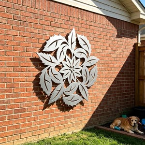 May include: A large, circular, silver-colored metal wall art piece featuring a symmetrical floral design with pointed, leaf-like petals radiating from a central star-shaped flower, mounted on a flat, red-brown brick exterior wall surface.