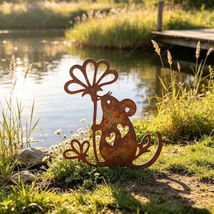 Op de afbeelding: Een roestkleurige metalen tuinsteker in de vorm van een muis die een grote bloem vasthoudt. Het silhouet van de muis heeft twee hartvormige uitsparingen op het lichaam en een lange gebogen staart, met een textuurrijke, verweerde afwerking.