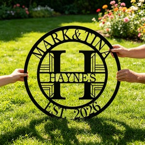 Puede incluir: Letrero de nombre familiar de metal negro con los nombres "Mark & Tina" y el apellido "Haynes" en el centro, con el año "EST. 2026" en la parte inferior. El letrero se sostiene sobre hierba verde.