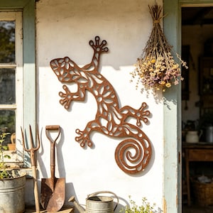 Puede incluir: Escultura de pared de metal oxidado con forma de lagarto y diseño floral. La pieza está montada en una pared blanca, cerca de una ventana y una puerta. Se ven herramientas de jardín y una regadera en primer plano.