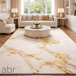 May include: A cream-coloured area rug featuring a gold leaf design. The rug is in a living room with a white sofa, armchairs, and a round coffee table. The room has a chandelier and large windows.