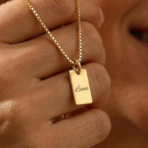 Puede incluir: Un collar con colgante rectangular dorado con el nombre "Luna" grabado. El colgante cuelga de una delicada cadena. El collar está siendo sostenido por una persona.