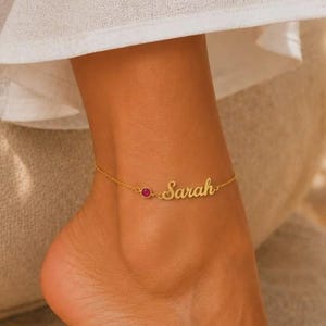 May include: A gold-toned anklet featuring a delicate chain, a small circular red gemstone, and a cursive nameplate with the name Sarah. The jewelry is worn around the ankle, showcasing a polished metal finish and a minimalist design aesthetic.