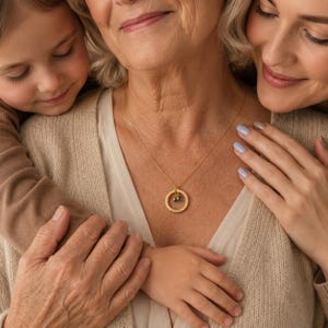 May include: A gold-toned necklace featuring a circular pendant with an engraved surface and a small, dangling charm. The pendant hangs from a delicate gold-toned chain, resting against a beige knit garment worn by an individual.