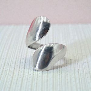 May include: A silver-toned ring with a unique, flowing design. The ring features two rounded, leaf-like shapes connected by a swirling band. The metal has a polished, reflective surface, creating a modern and elegant look. This piece is a statement accessory.