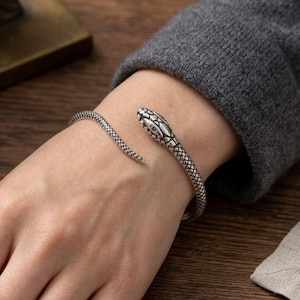 May include: A silver-toned metal cuff bracelet shaped like a coiled snake. The bracelet features a textured, scale-like pattern along the entire length of the body, with a detailed head design at one end and a tapered tail at the other end.