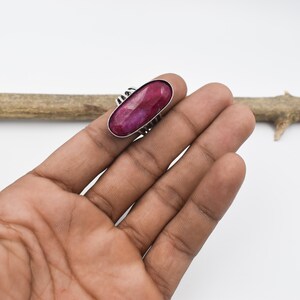 May include: A silver ring with a large, oval-shaped, deep red gemstone. The ring is set on a hand against a white background. The gemstone is the focal point, showcasing its rich color and polished surface.