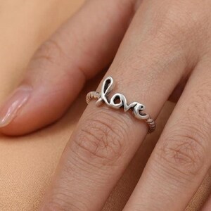 May include: A silver ring spelling out the word "love" in a cursive font. The ring band has a twisted rope design. The ring is worn on a finger against a neutral background.