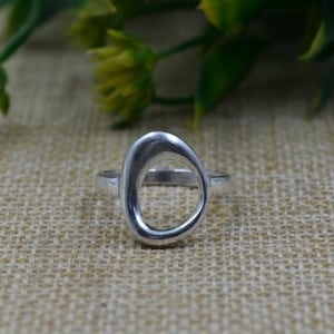 Puede incluir: Un anillo de plata con un diseño en forma de óvalo. El anillo está hecho de un material metálico brillante y se encuentra sobre una superficie de color neutro. El diseño del anillo es una forma ovalada lisa y abierta.