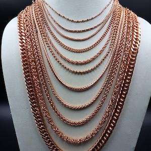 Può includere: Una collezione di dieci catene in metallo color oro rosa con vari spessori e stili di maglia, inclusi motivi a catena gourmette, a corda, a palline e a grano, sovrapposte su un busto espositore bianco per mostrare le diverse texture.