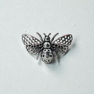 May include: A silver-colored bee-shaped brooch. The brooch features intricate cut-out details on the wings and body, with a textured surface. The wings are open, and the overall design is detailed and realistic.