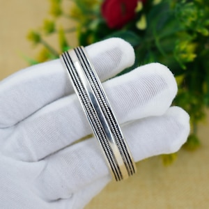 Puede incluir: Un brazalete plateado con una banda central pulida y bordes texturizados detallados. El brazalete está sostenido por una mano enguantada, con un fondo borroso de follaje verde y una flor roja.