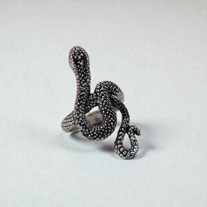 May include: A silver snake ring with a detailed, textured pattern. The snake is coiled, with its head raised. The ring is a unique piece of jewellery, likely made of metal, and would make a striking accessory.