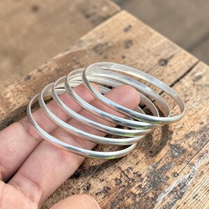 May include: Five silver bangles with a smooth, rounded design. The bangles are held in a hand, with a wooden surface in the background. The bangles are a bright, shiny silver colour.