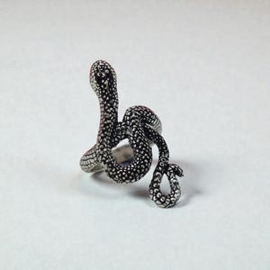 May include: A silver snake ring with a detailed, textured pattern. The snake is coiled, with its head raised, and the body wraps around the finger. The ring has a polished finish and is set against a white backdrop.